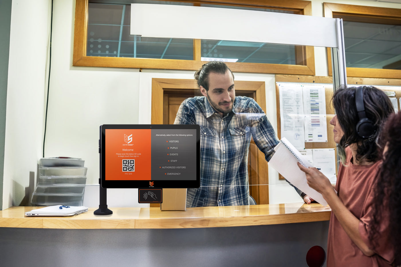 A man in a blue plaid shirt stands behind a service counter interacting with a customer wearing headphones and holding papers. A touchscreen terminal with an orange and black display is visible on the counter.