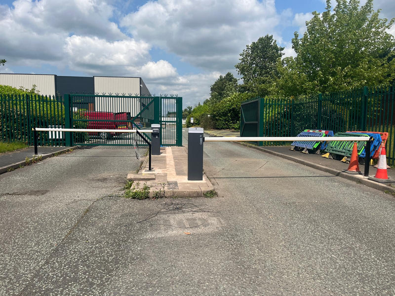 Came barrier gate at an entrance with traffic cones and a green fence.