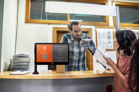 A man in a blue plaid shirt stands behind a service counter interacting with a customer wearing headphones and holding papers. A touchscreen terminal with an orange and black display is visible on the counter.