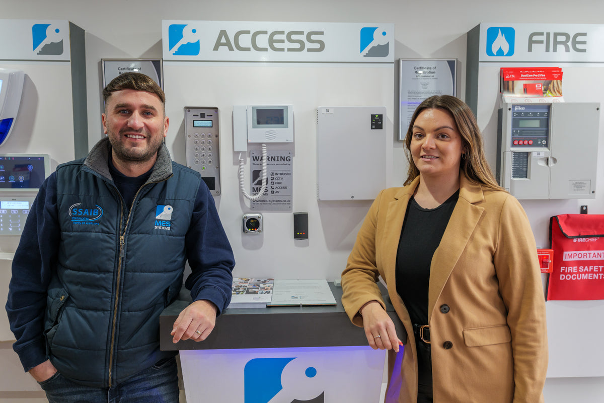A man in a MES Systems navy blue uniform jacket and a woman in a tan blazer stand behind a counter in a showroom displaying fire safety equipment, including Access and Fire alarm systems.