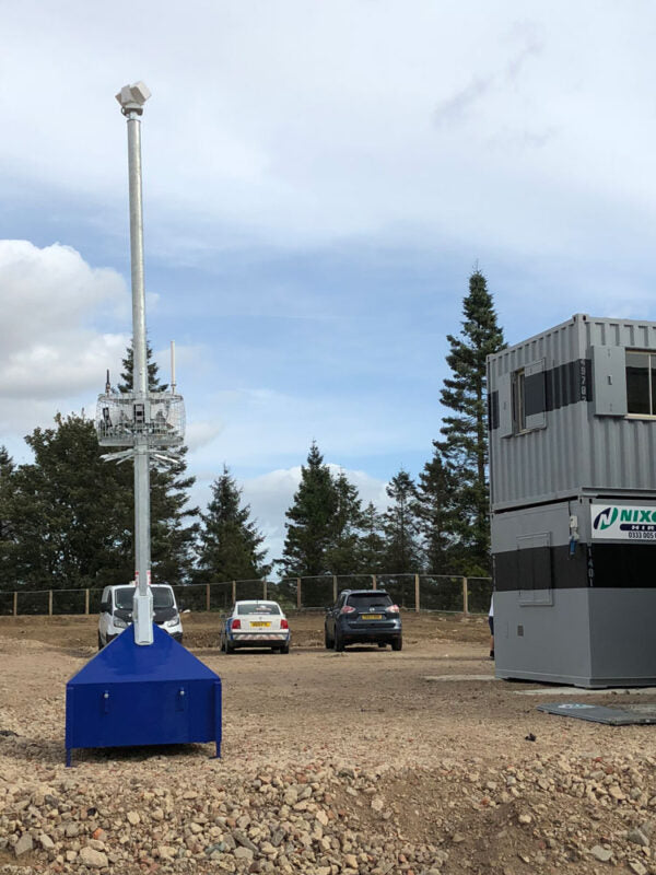 A large outdoor CCTV on a blue base next to a gray modular building, with cars and trees in the background under a cloudy sky.