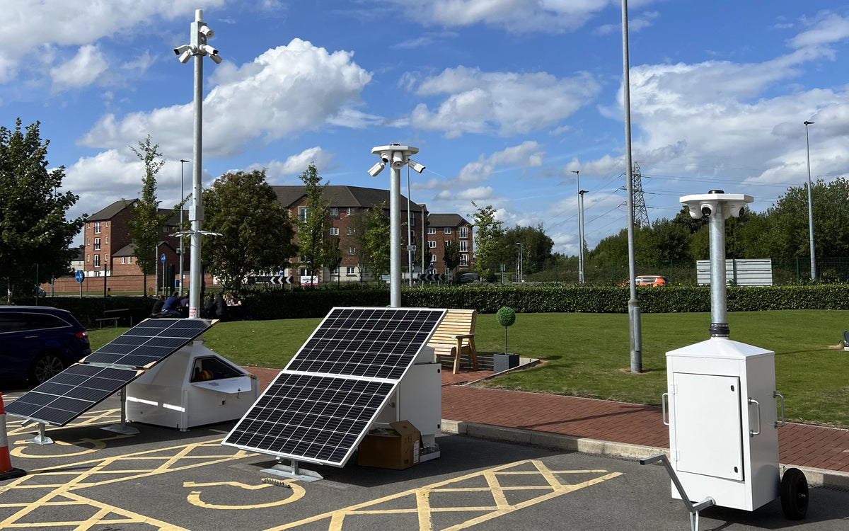 solar-powered CCTV equipment, including two solar panels on angled mounts and a white wheeled control unit, set in a park-like area with grass, trees, and buildings under a blue sky.