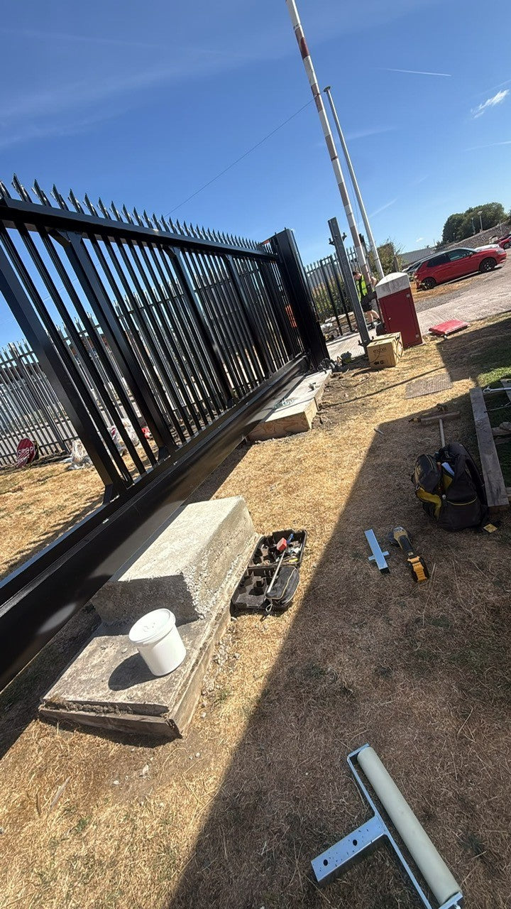 A black metal fence panel system installed outdoors on a dry grassy lot, with tools, concrete blocks, a white bucket, and construction equipment visible on the ground under a blue sky.