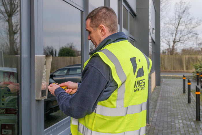 Person in a high-visibility vest installing access control, with a building and outdoor area in the background.