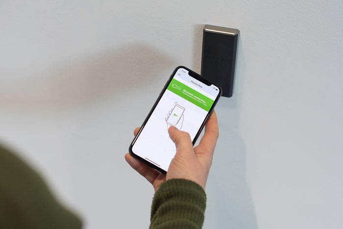 Hand holding a smartphone with an app displayed next to a wall-mounted device on a light gray background