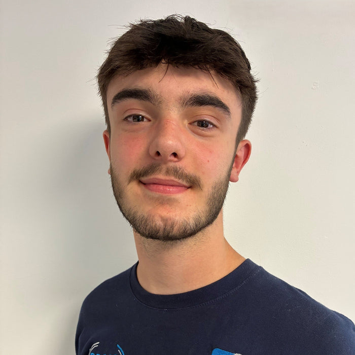 A young man with short brown hair and a light beard is wearing a navy blue t-shirt with 'SSAIB' and 'MES SYSTEMS' logos on the chest, photographed against a plain off-white wall.