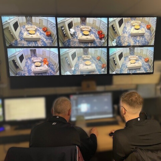 Two people monitoring multiple security camera screens displaying a room with furniture and decor.