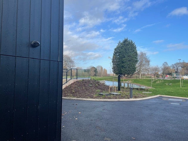 Black wooden-clad building with a discreet, black MES Systems CCTV camera and a view of a park and blue sky.