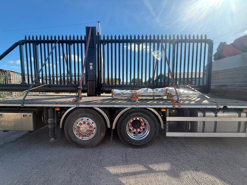 A black metal flatbed trailer with a large black gate mounted on the back, parked outdoors on a paved surface under a blue sky.