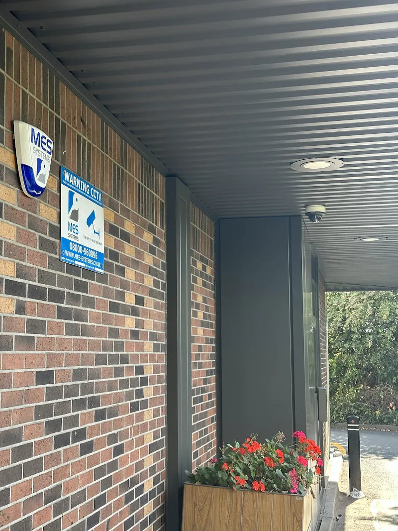 A McDonald's brick building exterior with a corrugated metal roof, blue and white MES warning signs on the wall, a wooden planter box with red and pink flowers, and a security camera mounted on the ceiling.