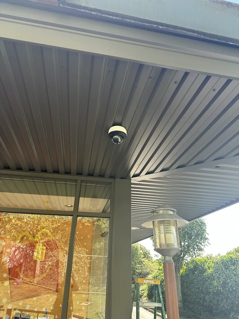 A dome security camera mounted on a corrugated metal roof overhang above a glass door at a McDonald's restaurant, with a garden light and outdoor greenery visible in the background.