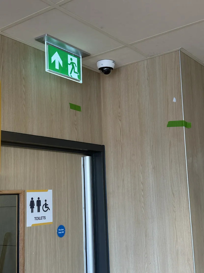 An interior hallway corner with light wood-paneled walls, a green illuminated emergency exit sign above a doorway, a dome security camera on the ceiling, and a toilet sign on the door.