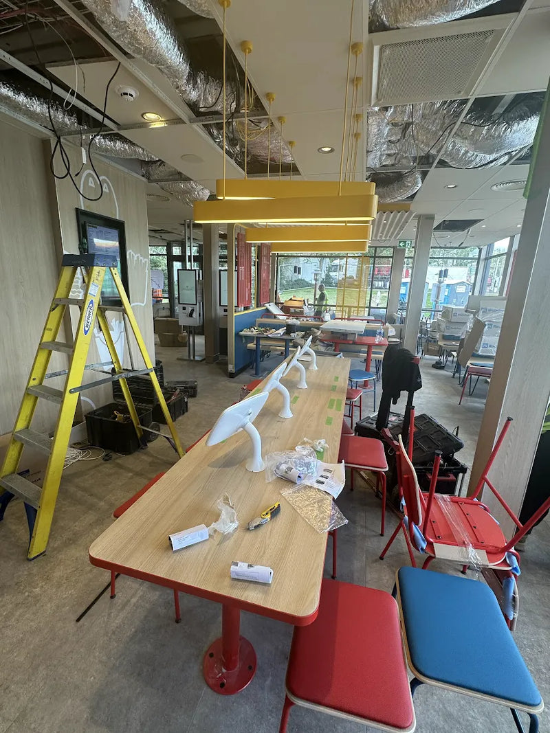 A long rectangular wooden table with red metal legs and matching red-framed chairs, shown in a restaurant interior under construction. With yellow pendant lights, exposed ceiling ducts, and construction equipment are visible around it.