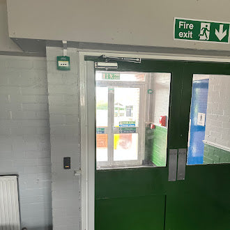 Green fire exit door at a school with Paxton Net 2 access control and a sign above it in a room with white tiled walls.
