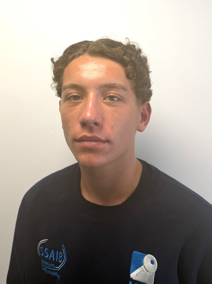 A young man with curly brown hair wearing a black crewneck sweatshirt with blue and white graphics on the chest, photographed against a plain off-white wall.