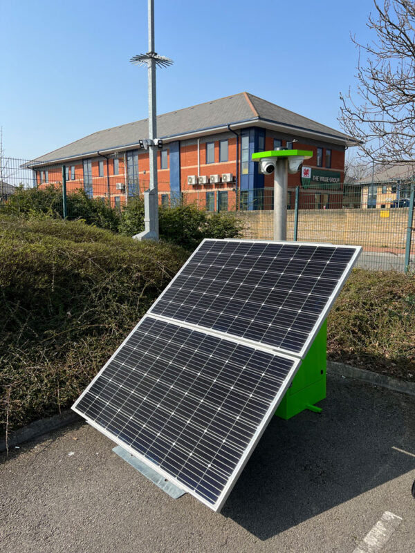 A solar panel for mobile CCTV mounted on a green metal stand outdoors, photographed in front of a brick building and fence under a clear blue sky.