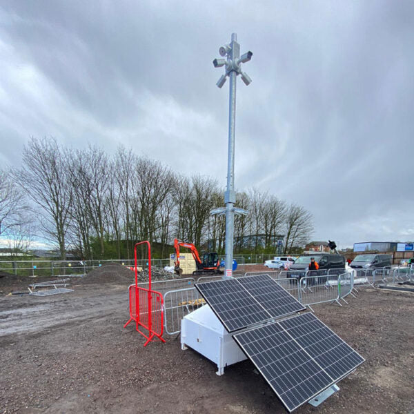 A portable solar power station with two angled solar panels and a tall metal light pole mounted on top, set in an outdoor construction or event site under a cloudy sky.