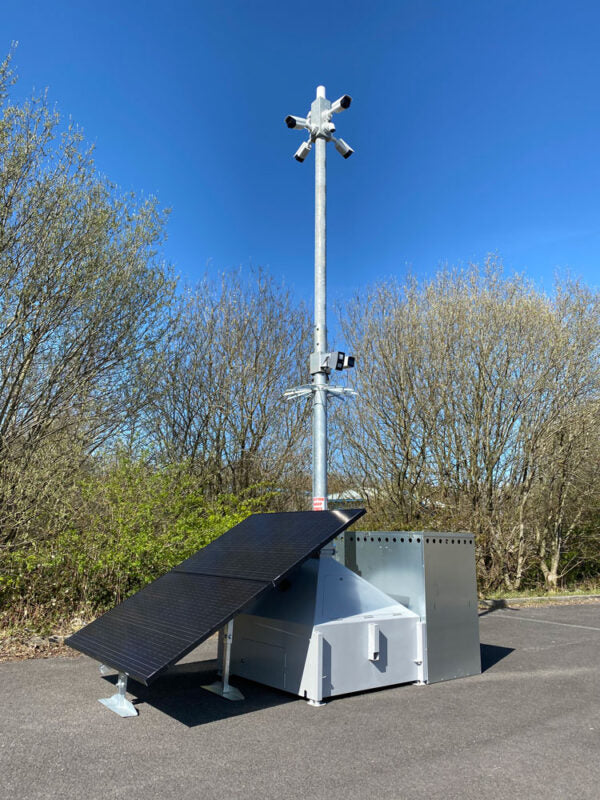 A solar-powered surveillance camera system with a tall metal pole, multiple cameras at the top, a tilted solar panel, and a gray metal base cabinet, photographed outdoors on a paved surface with trees and blue sky in the background.